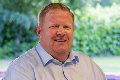 Professional headshot of a smiling man in a light blue shirt, photographed outdoors.